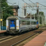 Une femme en fauteuil roulant devant une gare avec valise, assistée