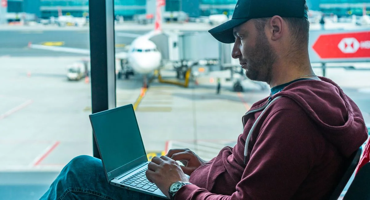 A man working on his laptop in an airport terminal, with a plane visible outside.