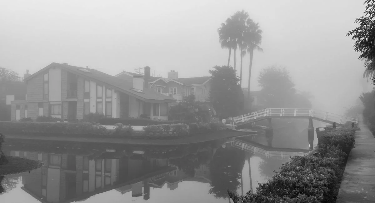 A tranquil foggy morning captures reflections of a bridge and houses in a canal.