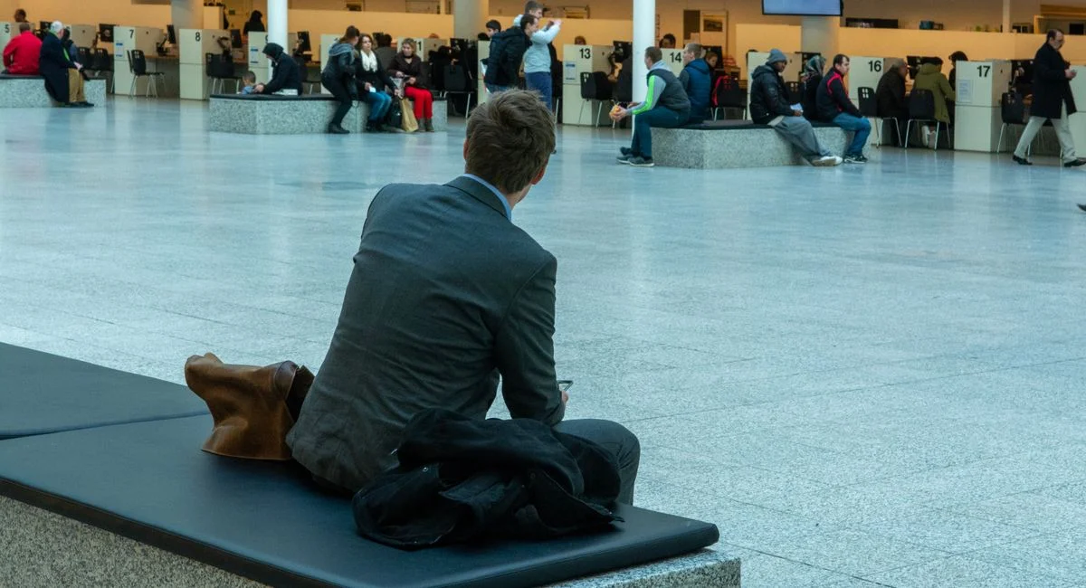 Businessman in suit seated in airport terminal waiting area, observing travelers.