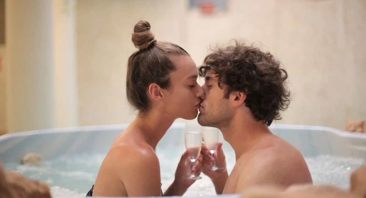 Side view of young tender man and woman sitting in bathtub with drinks in wine glasses and kissing each other enjoying weeken
