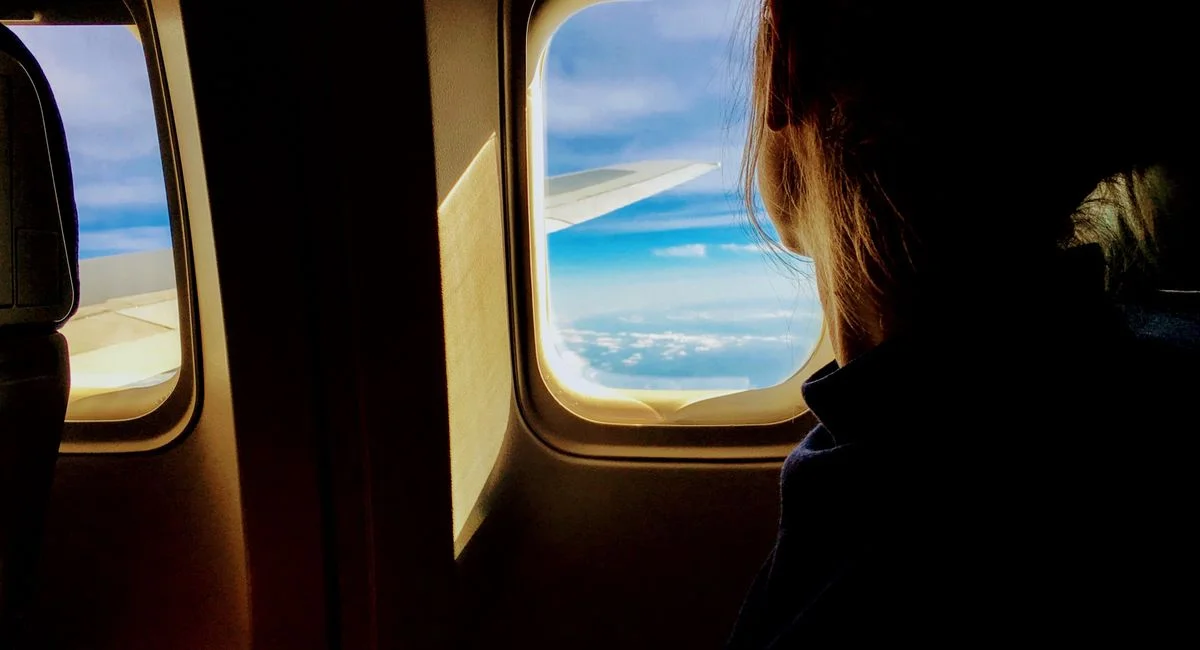 Silhouette of a woman looking out an airplane window at clouds and blue sky.