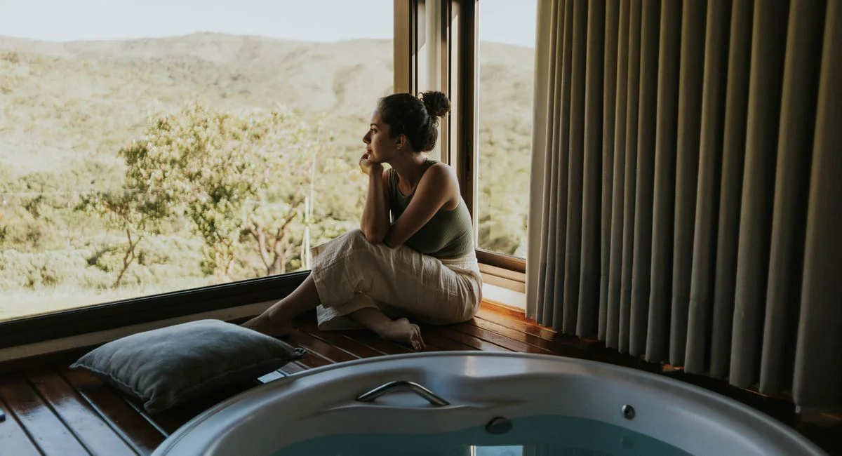 Woman gazing through window beside indoor jacuzzi overlooking mountains.